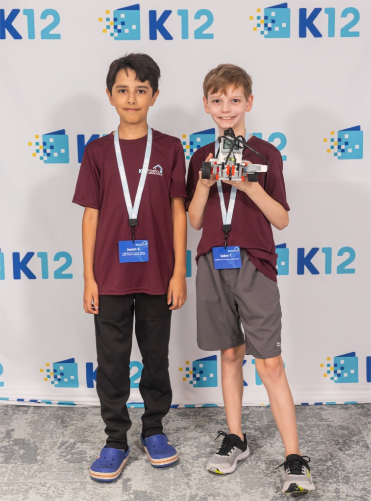 Dos niños con camisetas granates y etiquetas con sus nombres posan sonrientes delante de una pancarta de K12. Uno de ellos sostiene una maqueta de robot, que simboliza el trabajo en equipo y la innovación.