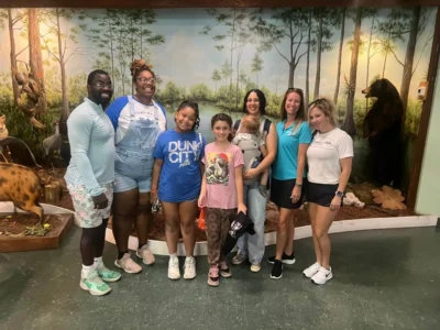 Siete personas, entre ellas dos niños, sonríen en el interior de un mural de temática forestal con figuras de animales.