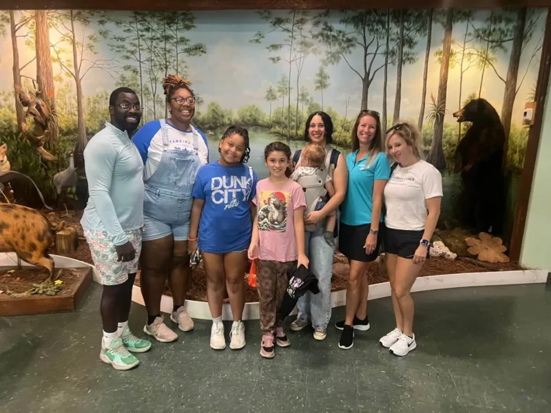 Siete personas, entre ellas dos niños, sonríen en el interior de un mural de temática forestal con figuras de animales.