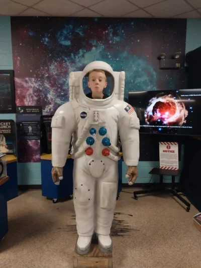 Un joven está de pie detrás de un gran astronauta, como si llevara puesto un traje espacial. La sala tiene paredes y expositores de temática cósmica, con un tono lúdico y educativo.