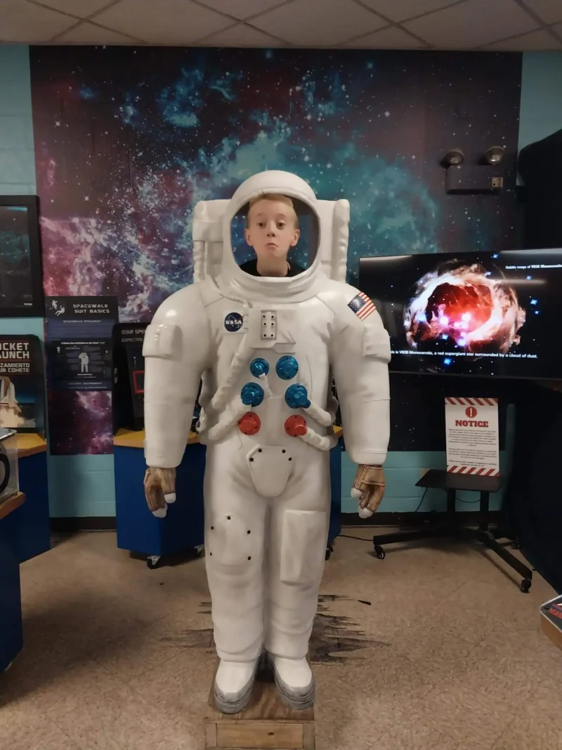 Un joven está de pie detrás de un gran astronauta, como si llevara puesto un traje espacial. La sala tiene paredes y expositores de temática cósmica, con un tono lúdico y educativo.