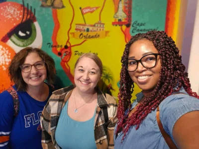 Tres mujeres sonríen delante de un colorido mural con mapas de lugares emblemáticos de Florida.